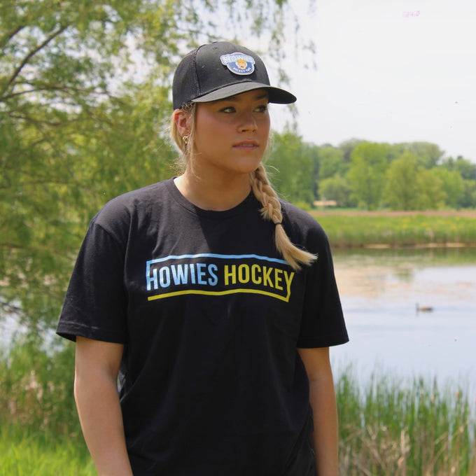 Person wearing a black 'Howies Hockey' t-shirt standing by a lake with trees in the background