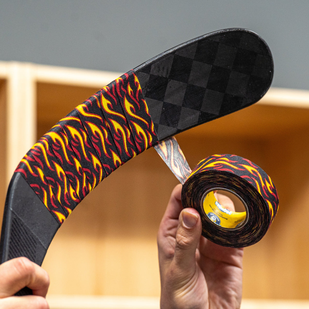 A person holds a hockey stick and a roll of hockey tape with a black, red, and yellow flame pattern in front of a wooden storage cubby wall.