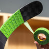A close-up of a hockey stick blade wrapped with bright green patterned tape, with a person holding a roll of the same tape in the foreground. The background is a softly blurred indoor setting with wood tones.