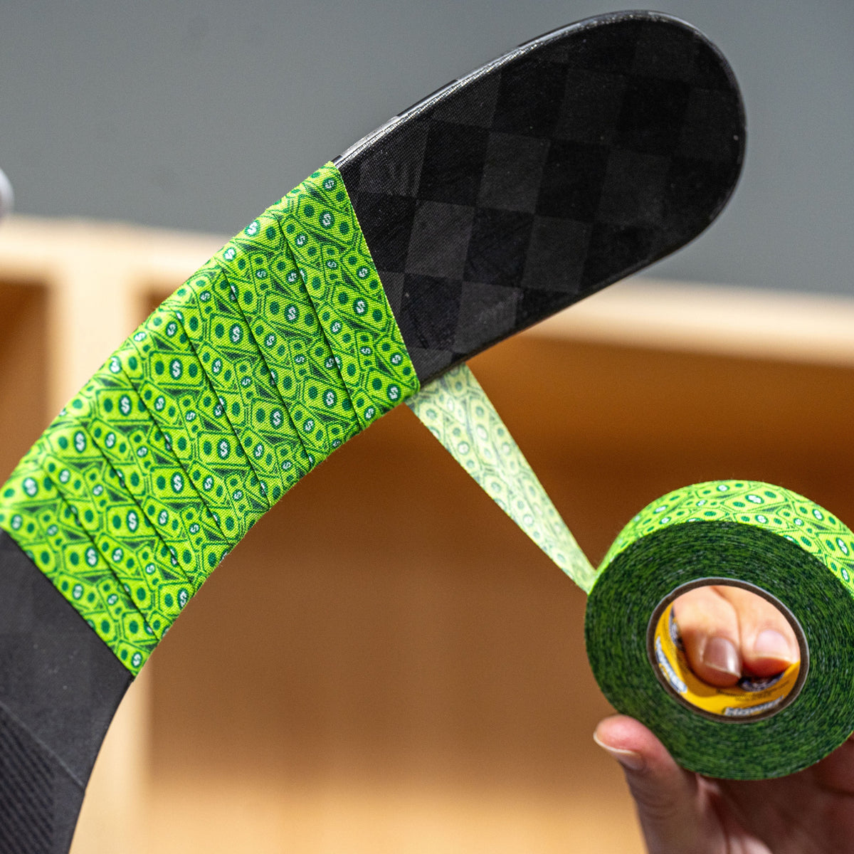 A close-up of a hockey stick blade wrapped with bright green patterned tape, with a person holding a roll of the same tape in the foreground. The background is a softly blurred indoor setting with wood tones.