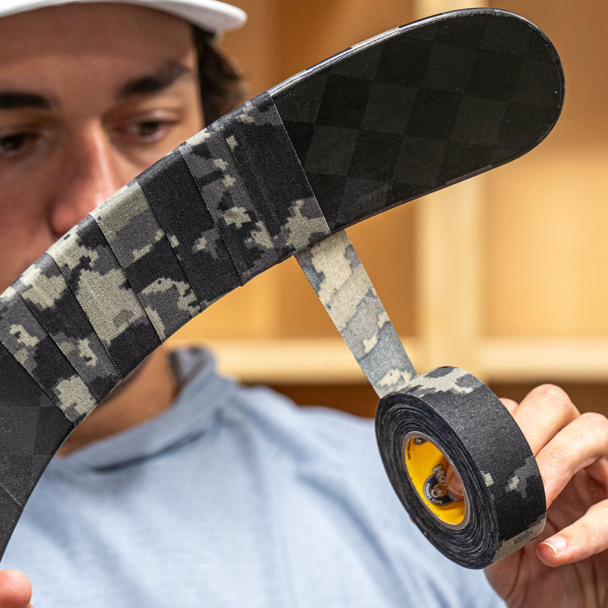 A man wearing a white cap with 'Howies' written on it holds up a roll of black Howies tape, showing both the black-and-white checkered unrolled section and the yellow core roll.
