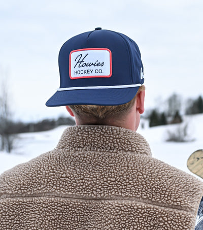 Person wearing a navy cap with a logo and holding a hockey stick in a snowy landscape