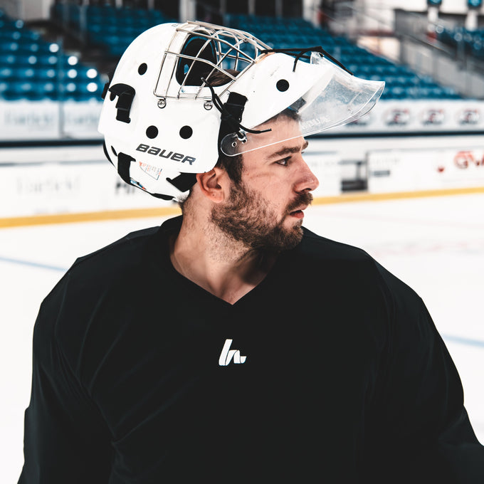 Person wearing a hockey goalie mask and blac jersey on an ice rink