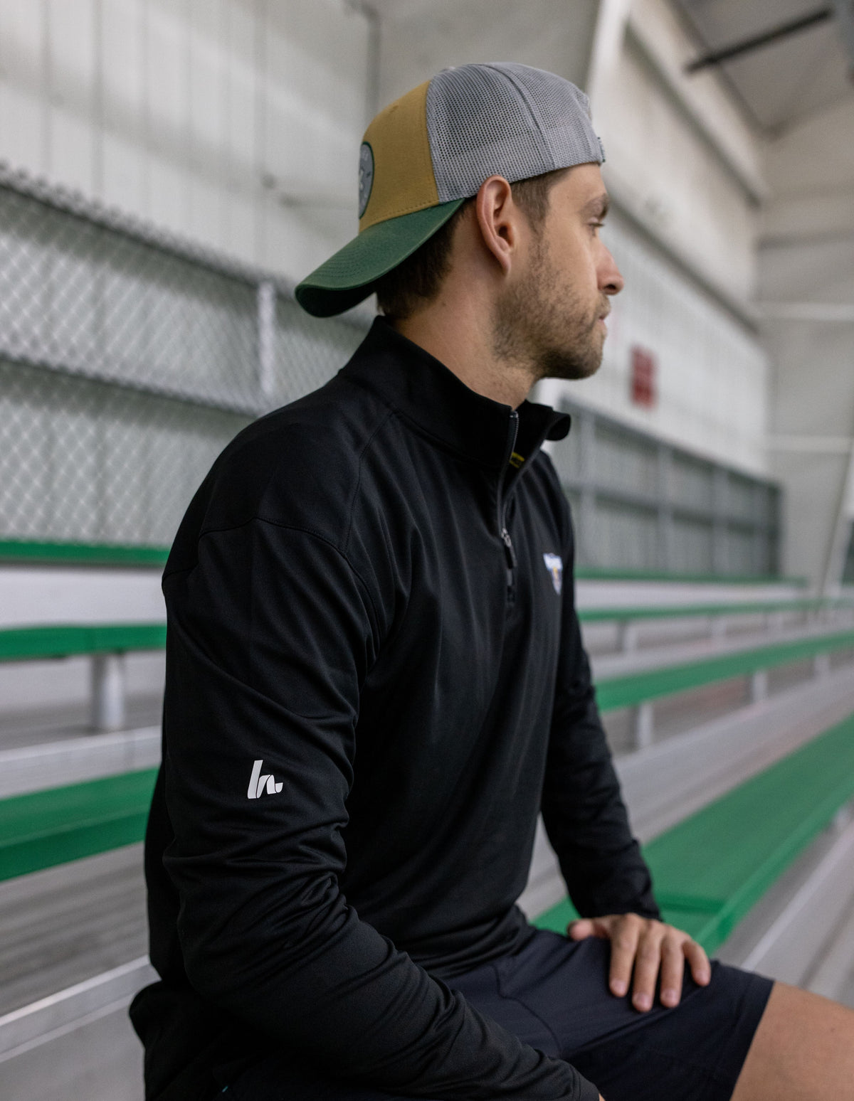 Man wearing a black jacket with a logo, sitting in a sports facility.