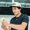 Hockey player wearing the Howies Clean Sheet Lid cream and black hat, smiling while unrolling white Howies hockey tape at the rink