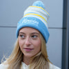 A woman wearing a light blue and white striped beanie with yellow stripes, featuring the text 'Howies HOCKEY CO' on the front and a blue pom-pom on top, set against a gray paneled wall.