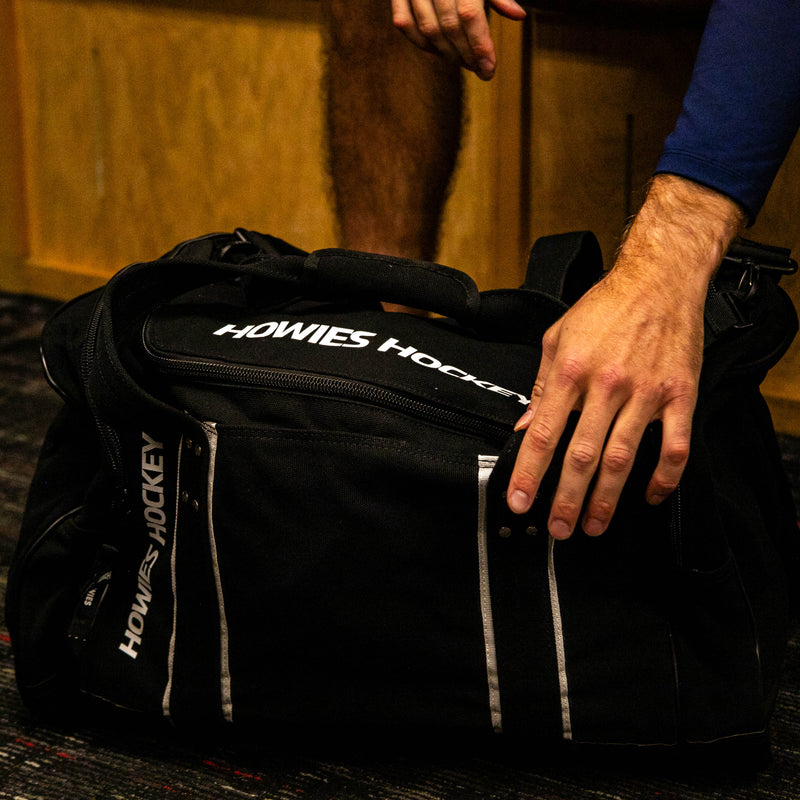 Person opening a black duffel bag with 'Howies Hockey' branding on a wooden floor.