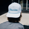 A person viewed from behind wearing a white baseball cap with 'Howies' embroidered in blue script on the front, standing outside near a building entrance.